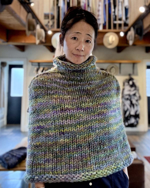 Person wearing a multicolored knitted poncho indoors with bookshelves in the background
