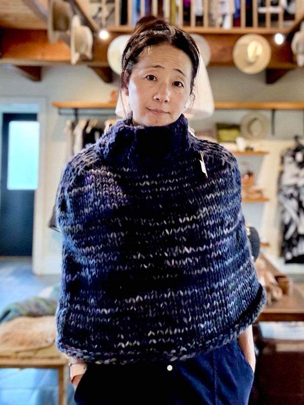 person wearing knitted turtleneck poncho in blue and purple gradient with wooden shelves in the background